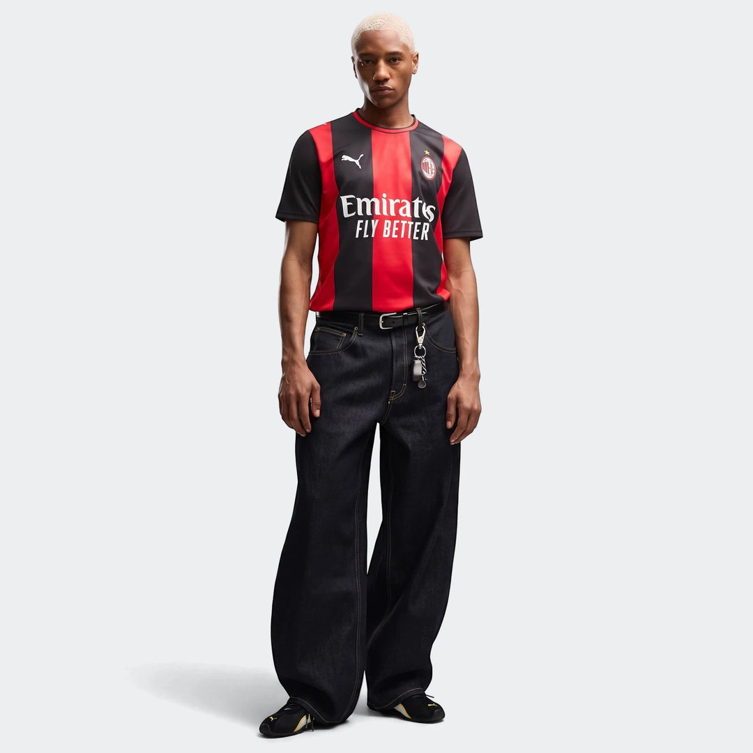 Full body shot of a male model wearing the black and red striped Ac Milan 26/27 Home Jersey, paired with dark wide-leg jeans
