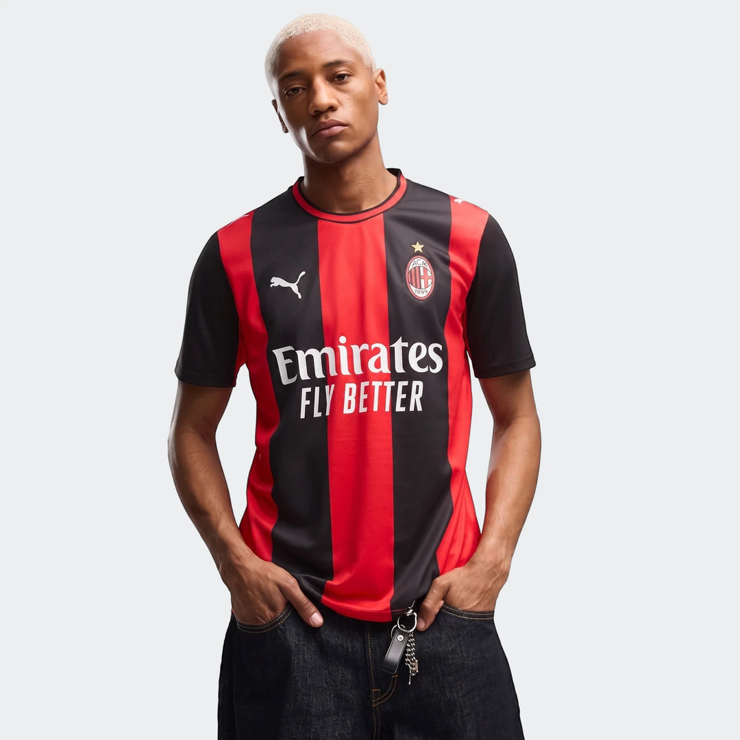 Male model wearing a red and black striped AC Milan 26/27 Home Jersey, standing with hands in pockets.