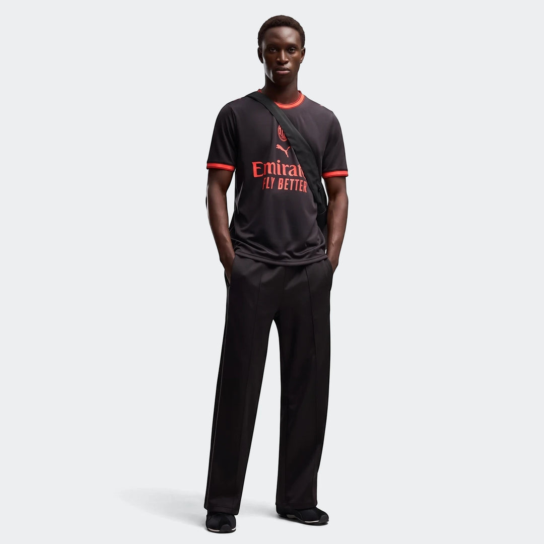 Male model wearing black AC Milan 26/27 Third Jersey with red accents and black pants, standing with hands in pockets.