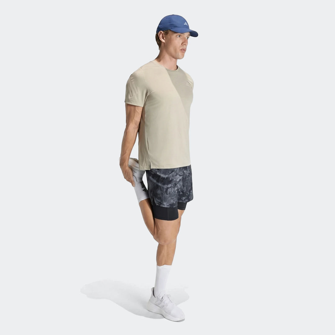 Male model in a beige Adidas Adi365 t-shirt, blue cap, and patterned shorts, stretching for a run.
