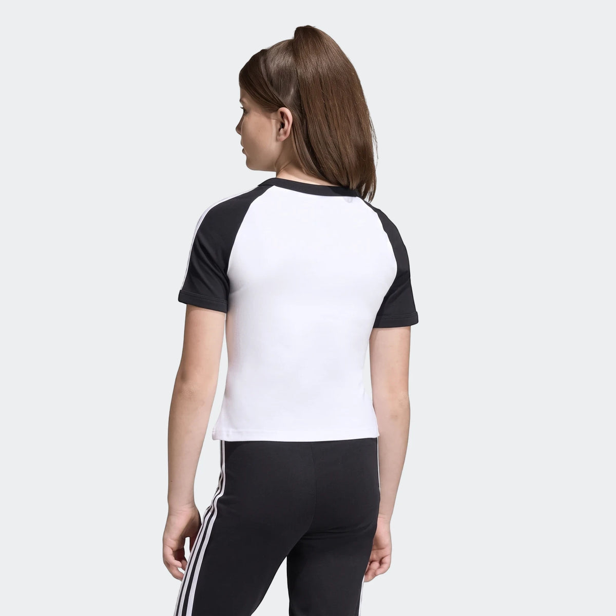 Young person from behind wearing a white and black Adidas 3 Stripes Slim T-Shirt and black striped pants.
