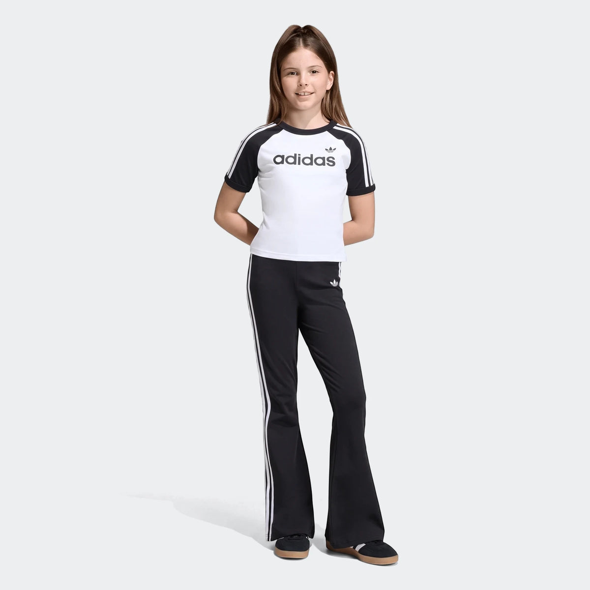 Young girl in a white Adidas 3 Stripes Slim T-Shirt with black raglan sleeves and black Adidas 3 Stripes track pants.