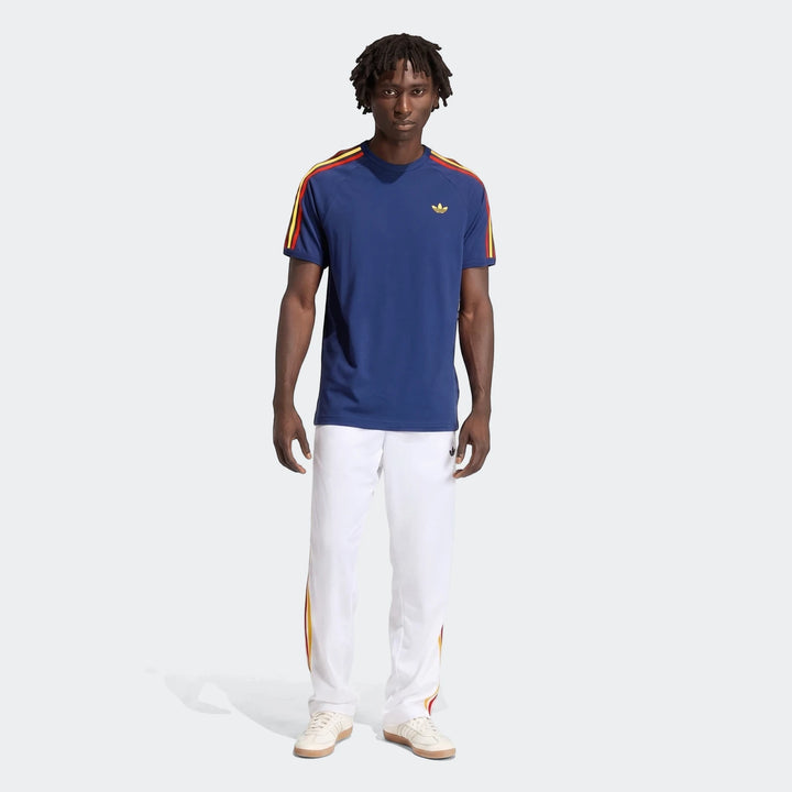 Male model wearing a navy blue Adidas 3-Stripes T-Shirt with red and yellow stripes and white track pants, shown in a
