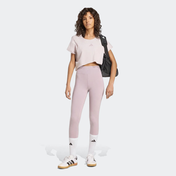 Model wears Adidas All Me Essentials Full-Length Tights in light purple with a pale pink crop top and white sneakers.