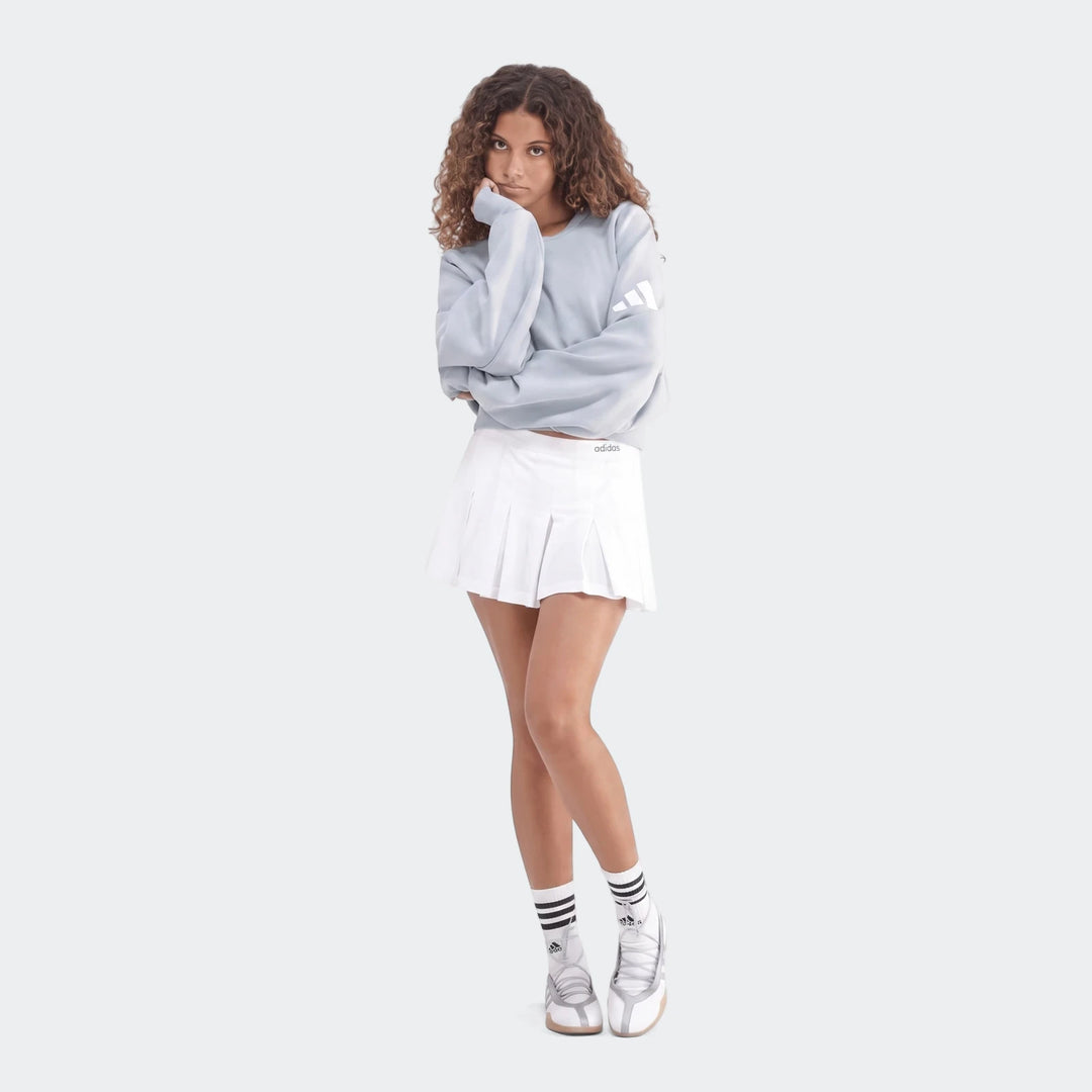 Young woman wearing a light blue Adidas All Season Slim Sweatshirt, white pleated tennis skirt, and white sneakers.