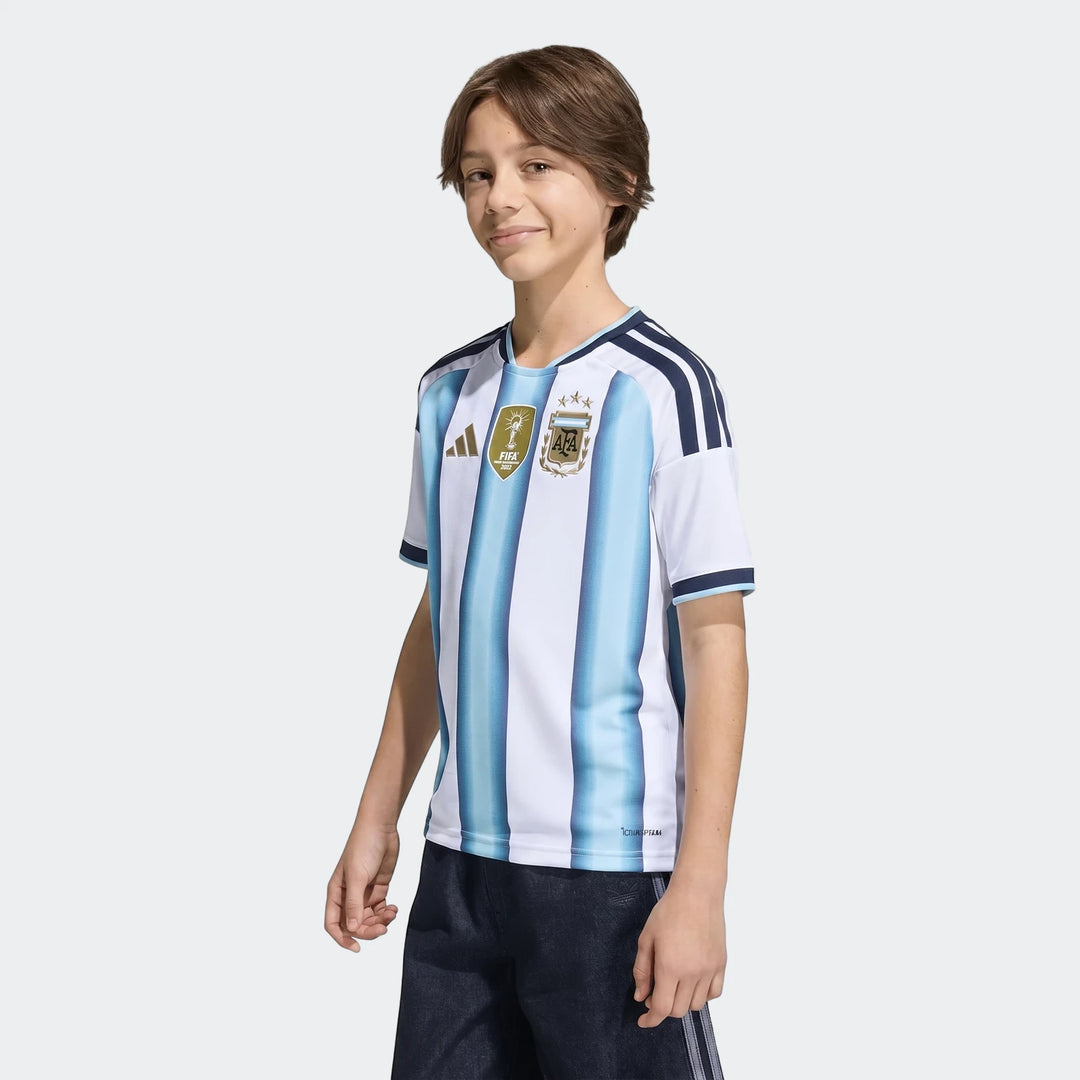 Young boy wearing the Adidas Argentina 26 Home Kids Jersey. Features white and light blue vertical stripes, short sleeves,