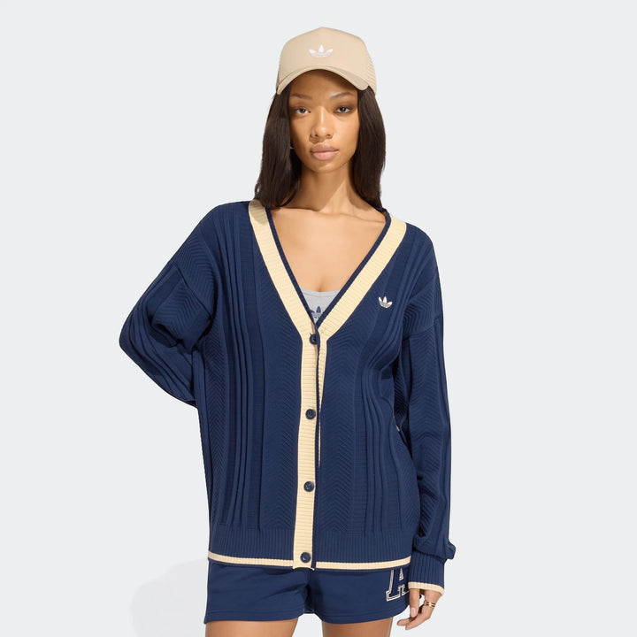A female model wearing a navy blue Adidas Cable Knit Cardigan, matching shorts, and a beige Adidas cap, posing confidently.