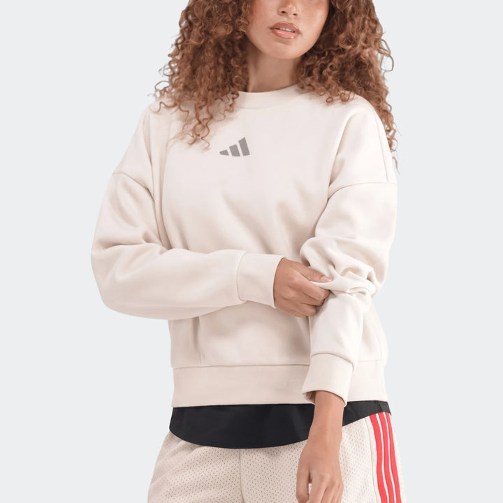 Woman wearing a cream Adidas Slim Fleece Crew Sweatshirt and mesh shorts, rolling up sleeve, front view.