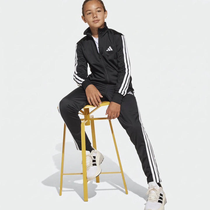 Young person wearing a black Adidas Essentials Climacool Track Suit with white stripes and white sneakers, sitting on a