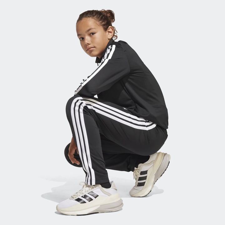 A young girl in a black Adidas Essentials Climacool track suit with white 3-stripes, crouching. The suit features a full-zip