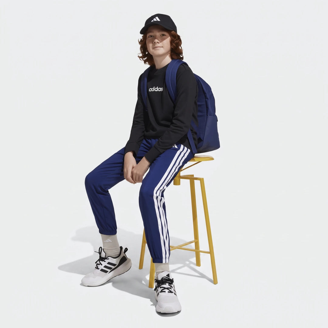 Young boy wearing Adidas Essentials Climacool blue tracksuit bottoms with white stripes, black sweatshirt, and a cap.