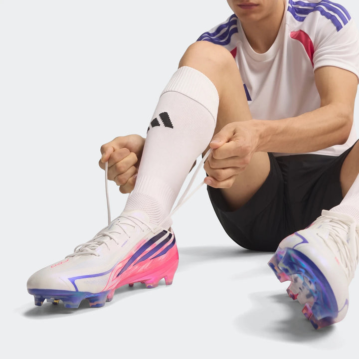 A person seated, tying the white laces of an Adidas F50 Hyperfast Elite Firm Ground Football Boot, featuring iridescent