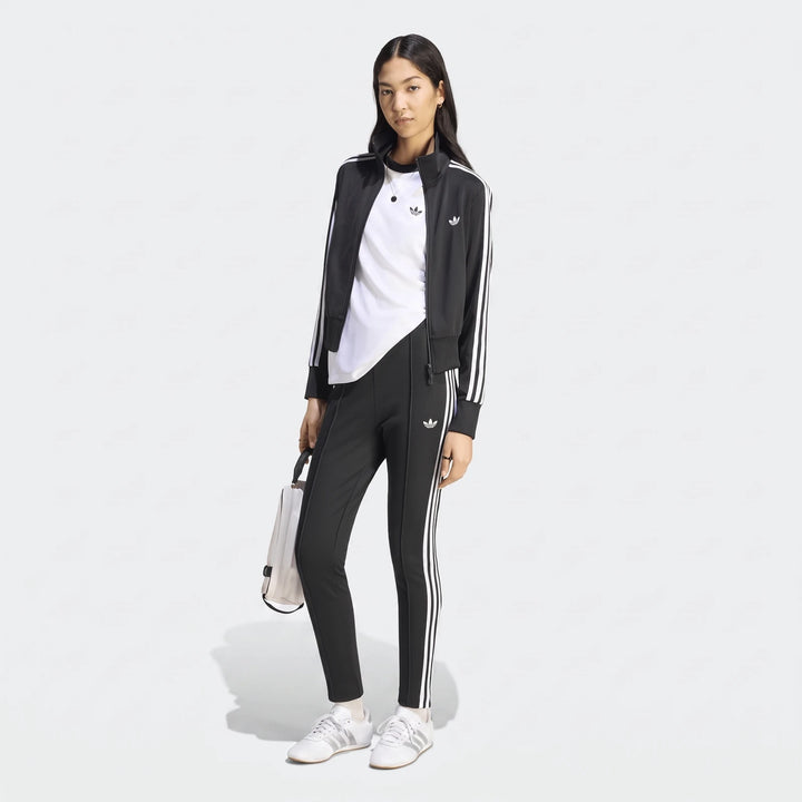 Female model in a black Adidas Firebird Classic Track Jacket with white stripes, matching track pants, white t-shirt, and