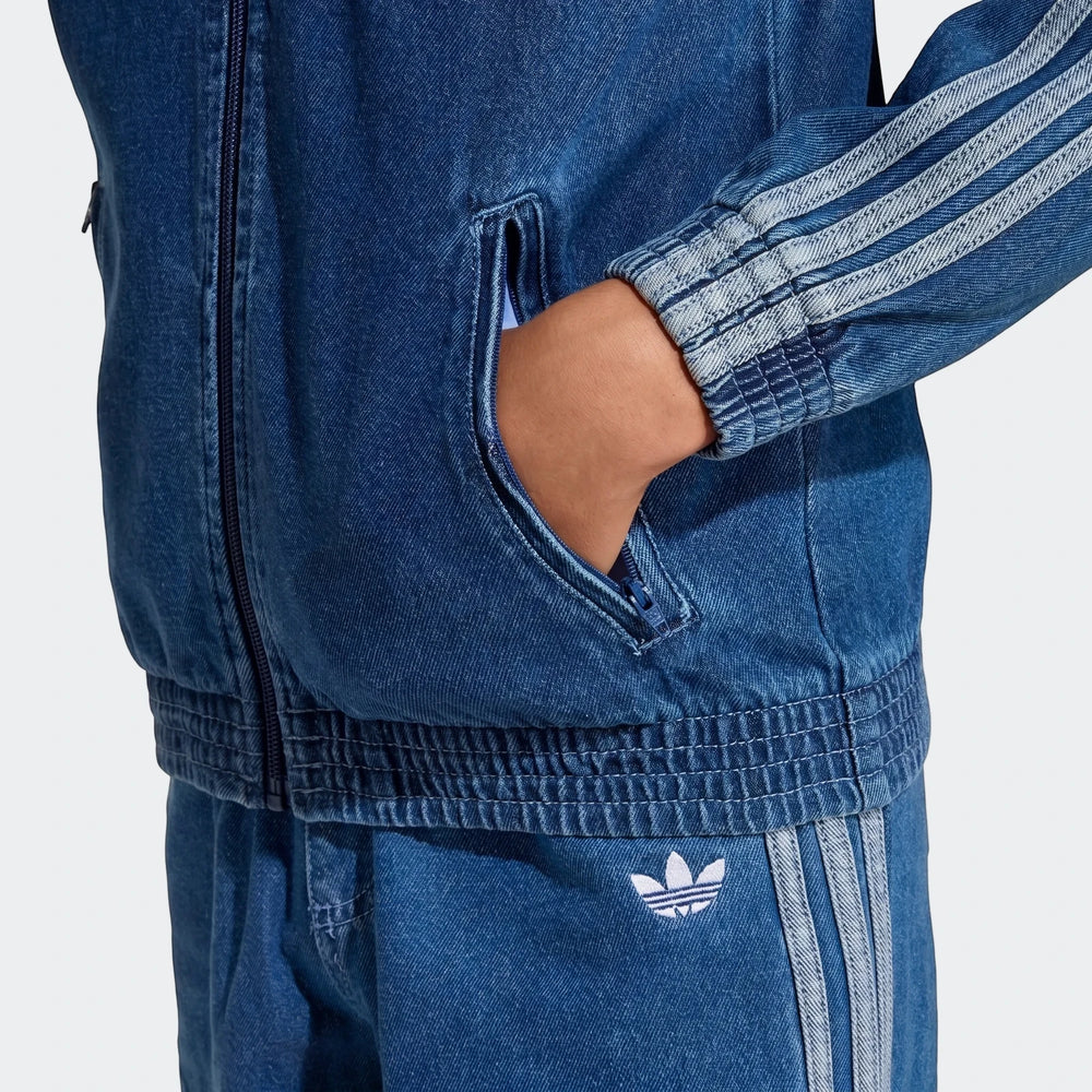 Close-up of a person wearing a blue adidas Firebird Denim Track Top, featuring a full zip and 3-stripes, with hand in pocket.