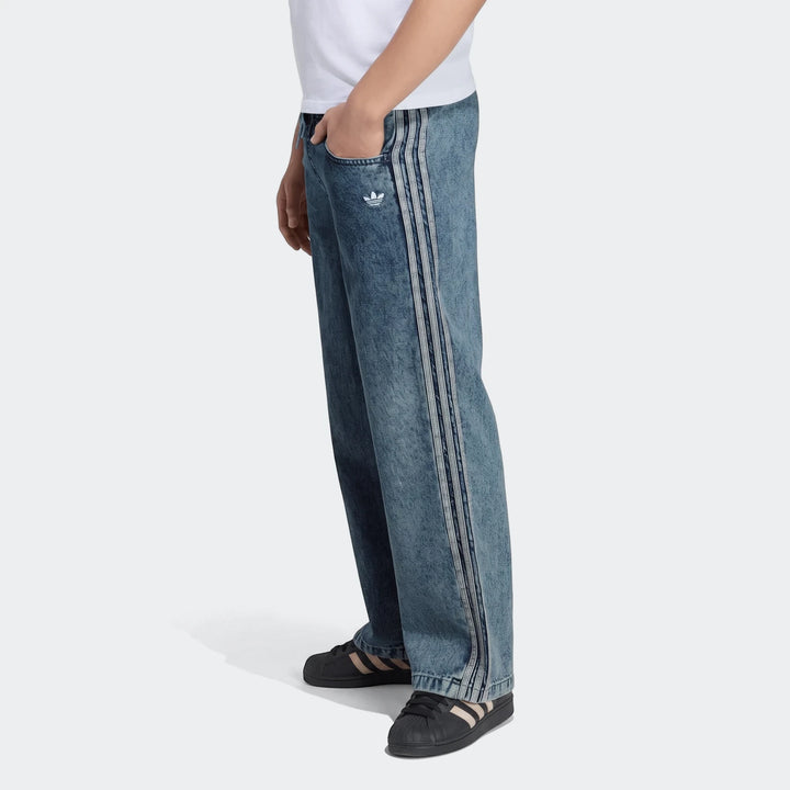 A person wears Adidas Firebird Loose Denim Track Pants in blue acid-wash with iconic three-stripes down the sides. Paired