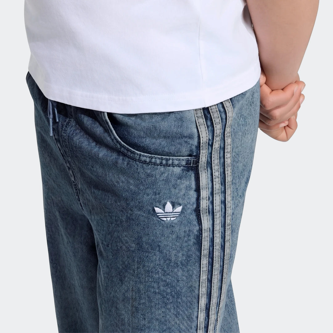 Close-up of blue Adidas Firebird Loose Denim Track Pants with three stripes and a Trefoil logo, worn by a person.