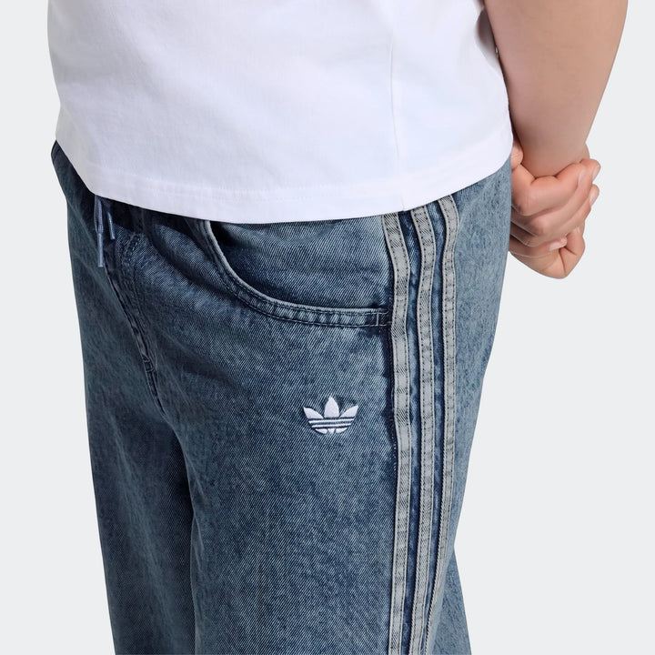 Close-up of blue Adidas Firebird Loose Denim Track Pants with three stripes and a Trefoil logo, worn by a person.