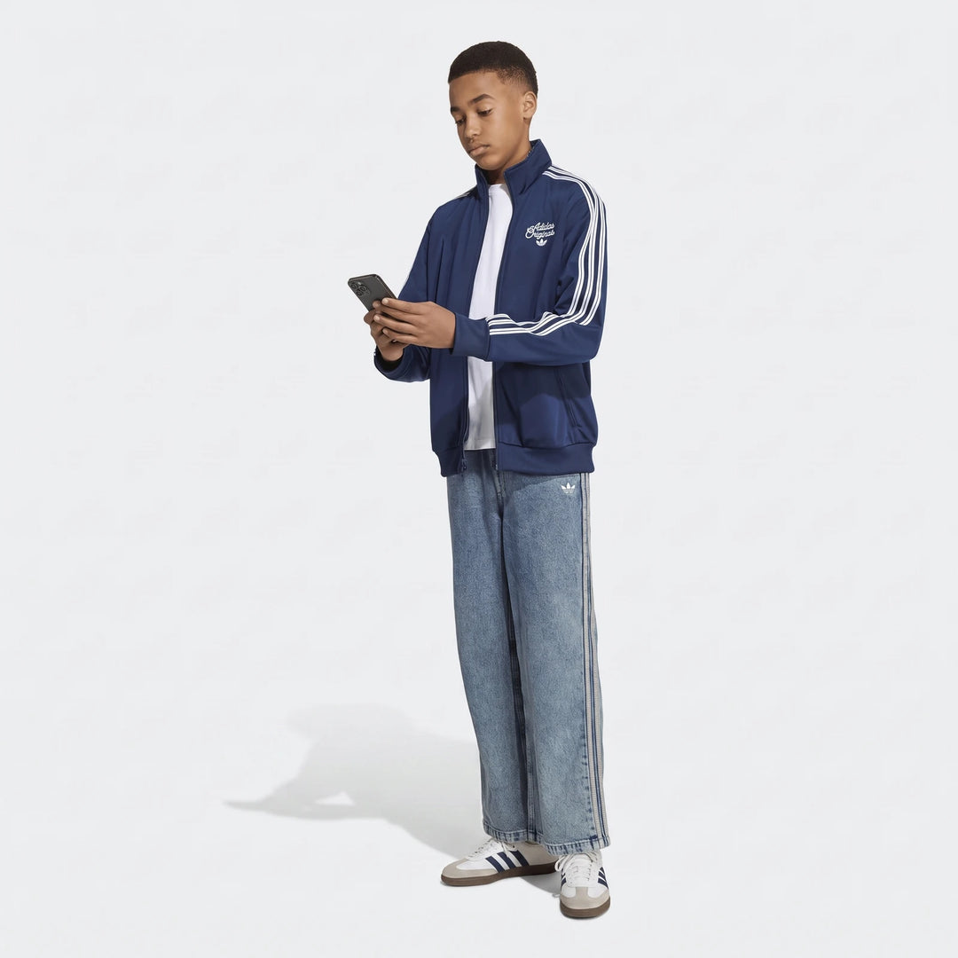 Young male model wearing a navy Adidas Firebird Loose Reversible Tracktop, light wash jeans, and sneakers, looking at a
