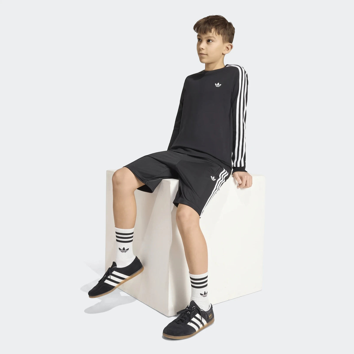 Boy wearing black Adidas Firebird shorts with white stripes, a black long-sleeve shirt, white socks, and black sneakers,