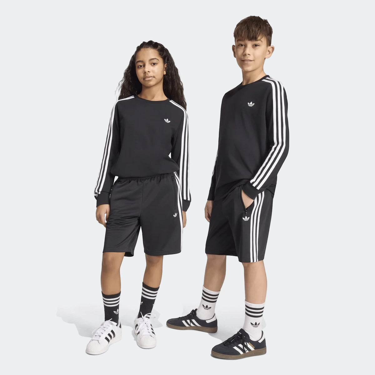 Two children model black Adidas Firebird shorts and matching black long-sleeve shirts with white stripes, paired with Adidas