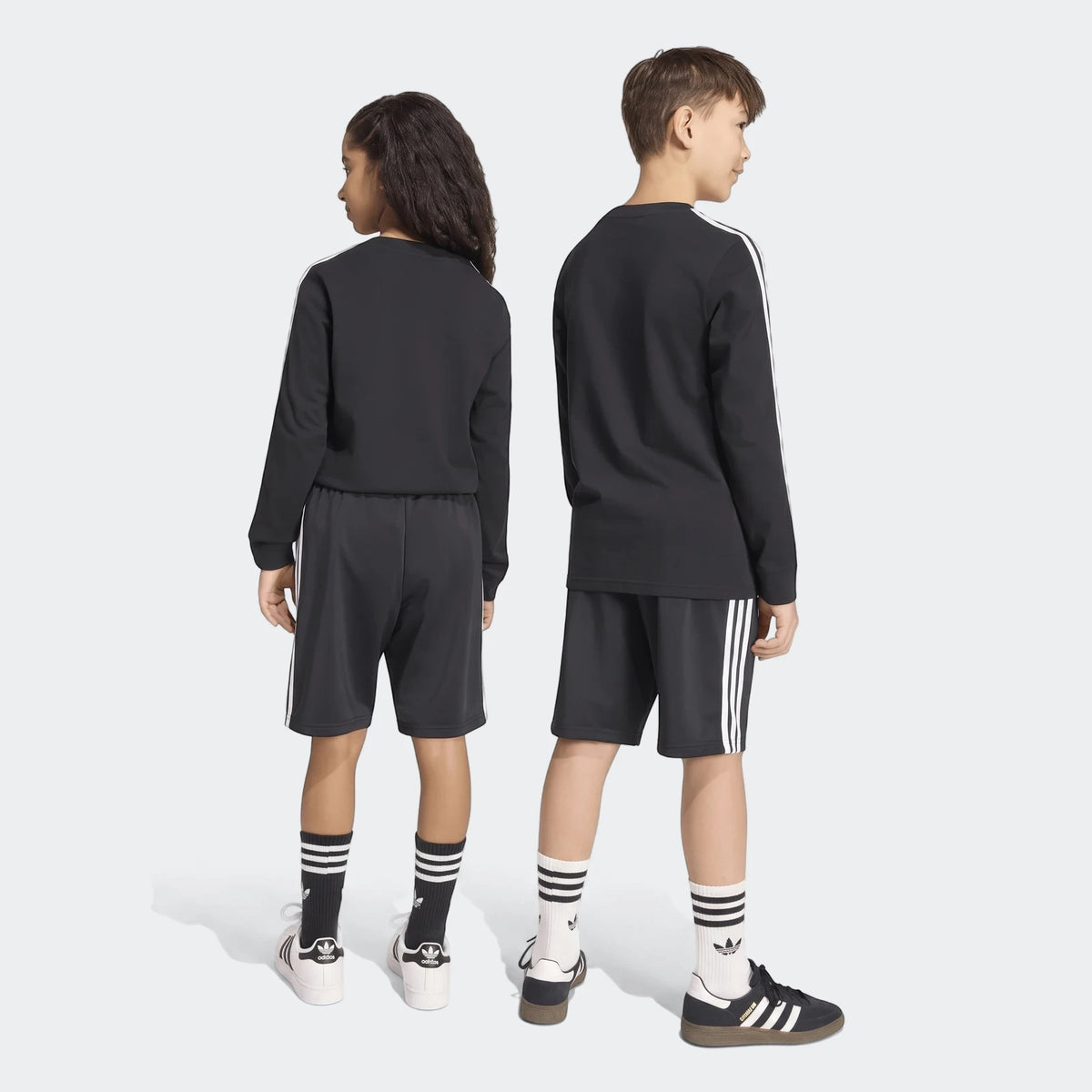Black Adidas Firebird Shorts and striped tops on a boy and girl, viewed from behind, with Adidas footwear.