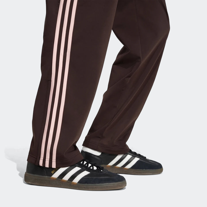 Dark brown Adidas Firebird track pants with light pink stripes on the side, worn with black and white Adidas sneakers,