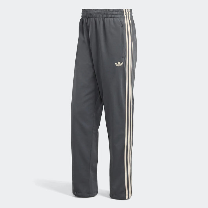 Dark gray Adidas Firebird Track Pants with off-white stripes and logo, designed for athletic wear, shown standing upright.