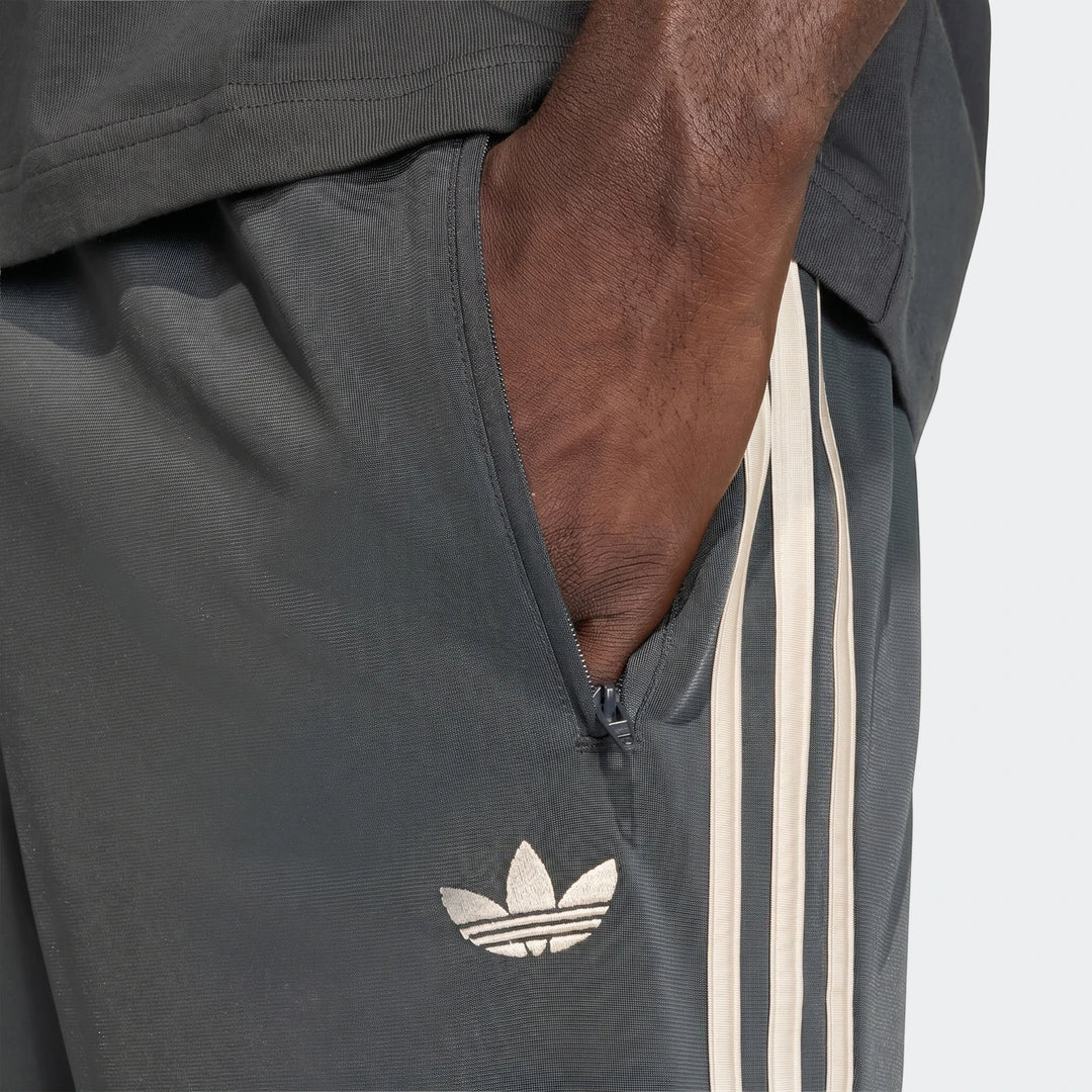 Close-up of dark grey Adidas Firebird Track Pants with off-white three stripes and a Trefoil logo, worn by a person.
