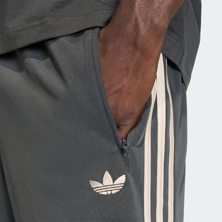 Close-up of dark grey Adidas Firebird Track Pants with off-white three stripes and a Trefoil logo, worn by a person.