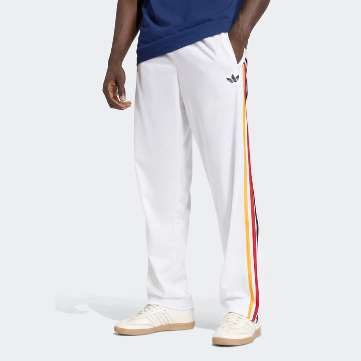 White Adidas Firebird Track Pants with red, yellow, and black side stripes, worn by a model from the waist down, showcasing