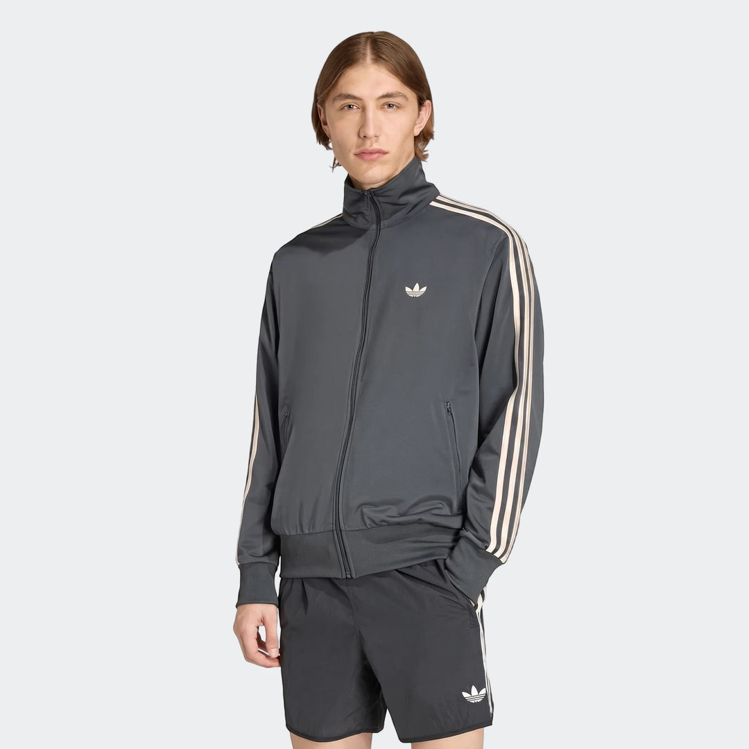 Male model wearing a dark grey Adidas Firebird track top and matching shorts, featuring signature three stripes and trefoil