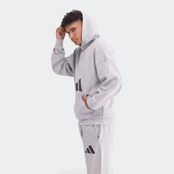 Young man modeling a light grey Adidas Future Icons 3-Bar hoodie and matching sweatpants set, comfortable and stylish.