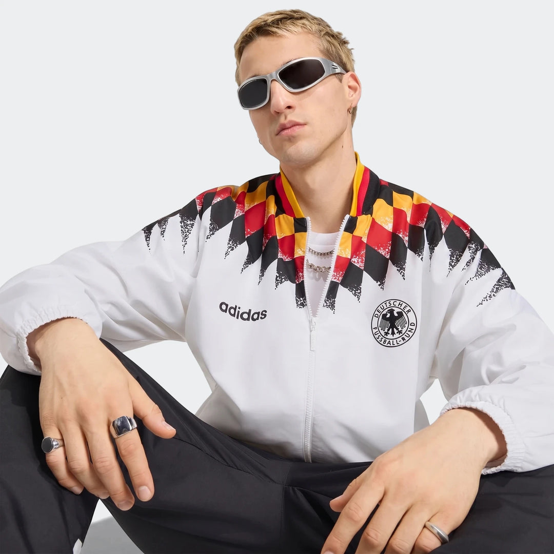Male model wearing a white Adidas Germany 1994 Retro Track Top with black pants and silver sunglasses, seated.