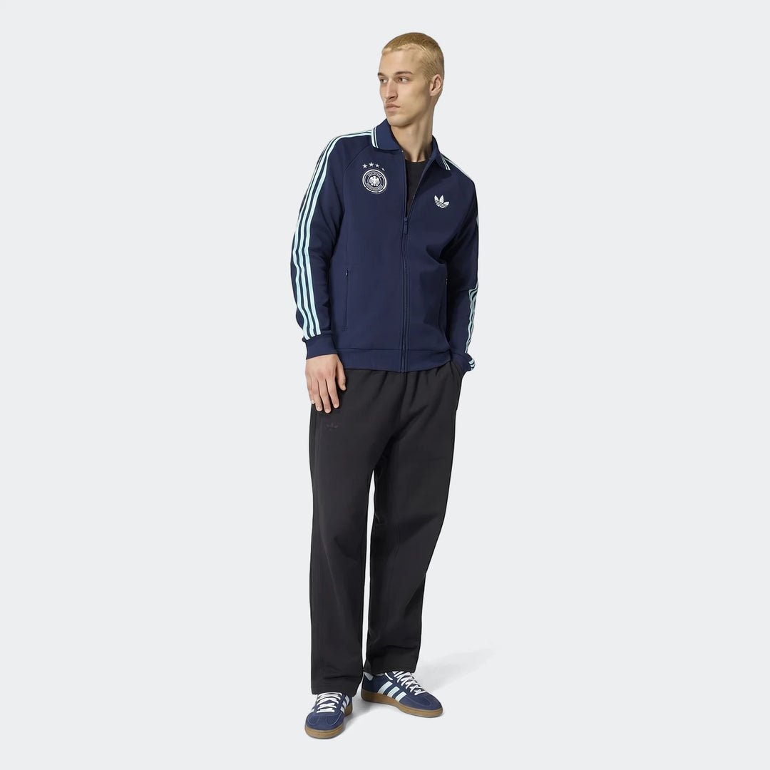 Young man wearing a navy blue Adidas Germany Away Originals track jacket, black sweatpants, and blue Gazelle sneakers.