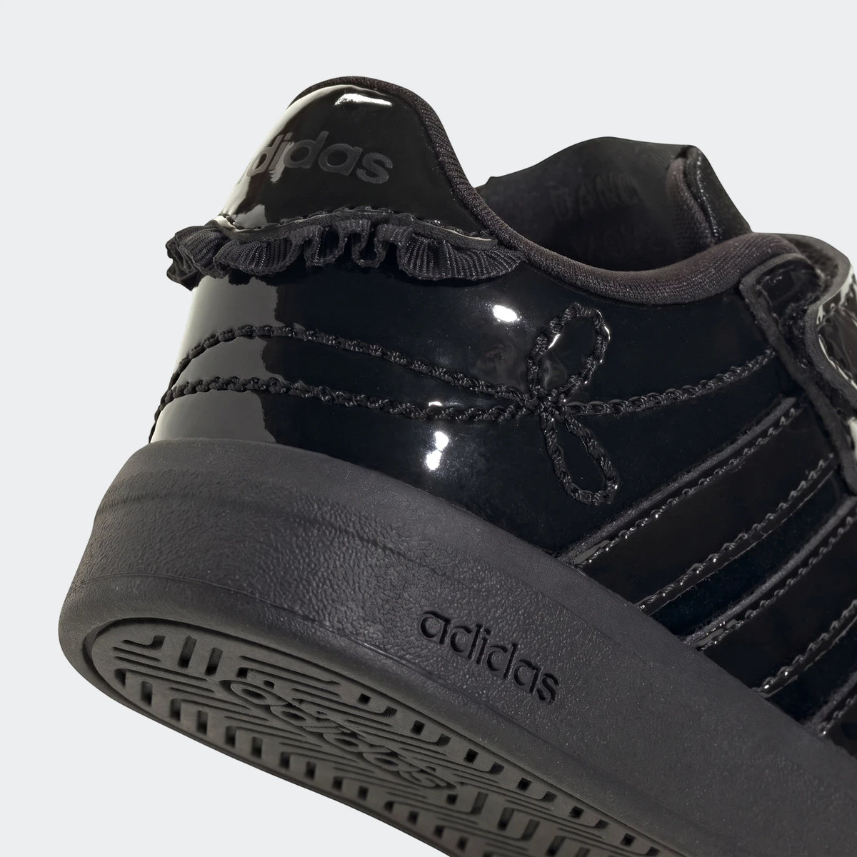 Close-up of a black Adidas Grand Court 3.0 infant girl's shoe, featuring patent leather, ruffled trim, and a visible sole.