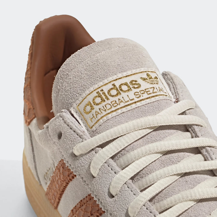 Close-up of a grey Adidas Handball Spezial shoe with brown textured stripes, white laces, and a gold logo on the tongue.