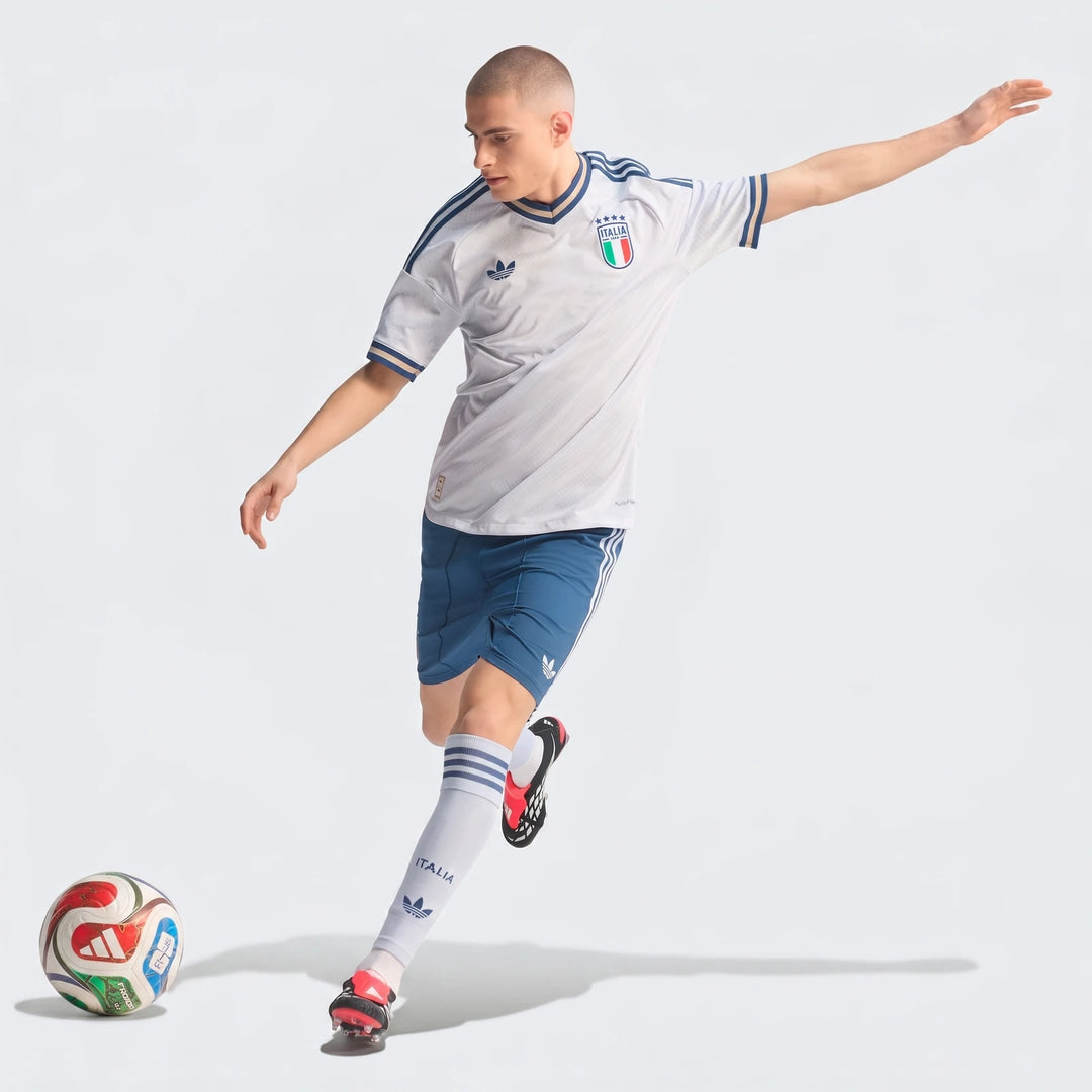 Male model in white Adidas Italia soccer jersey, blue shorts, and white socks, dribbling a soccer ball.