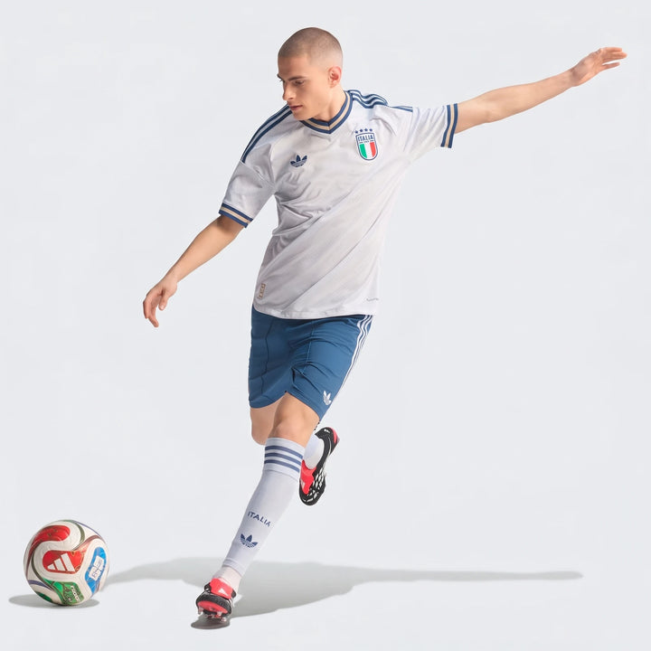 Male model in white Adidas Italia soccer jersey, blue shorts, and white socks, dribbling a soccer ball.