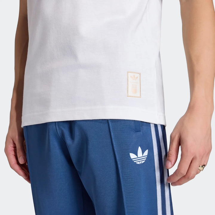 Close-up of a person wearing a white Adidas Italy Originals Graphic T-Shirt and blue track pants with white stripes,