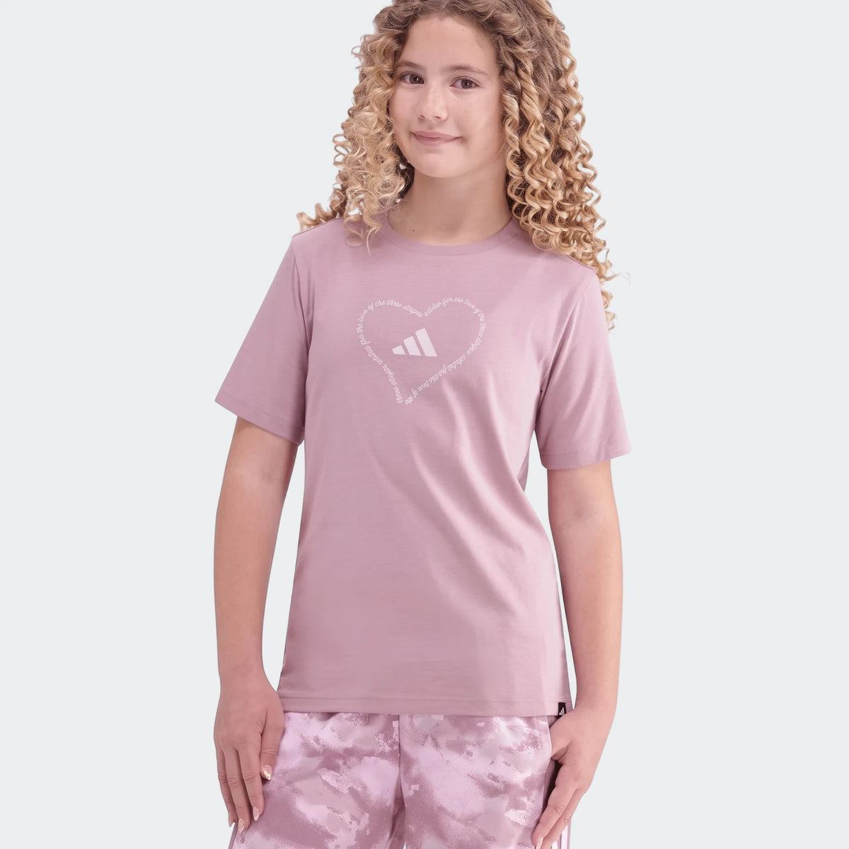 Young girl in a purple Adidas Jg Satin T-shirt featuring a white heart logo, paired with pink patterned athletic shorts.