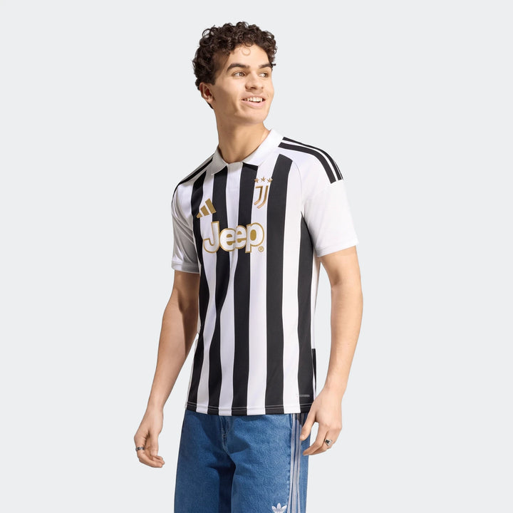 Smiling male model wears a black and white striped Adidas Juventus 26/27 Home Jersey with gold logos and blue jeans.