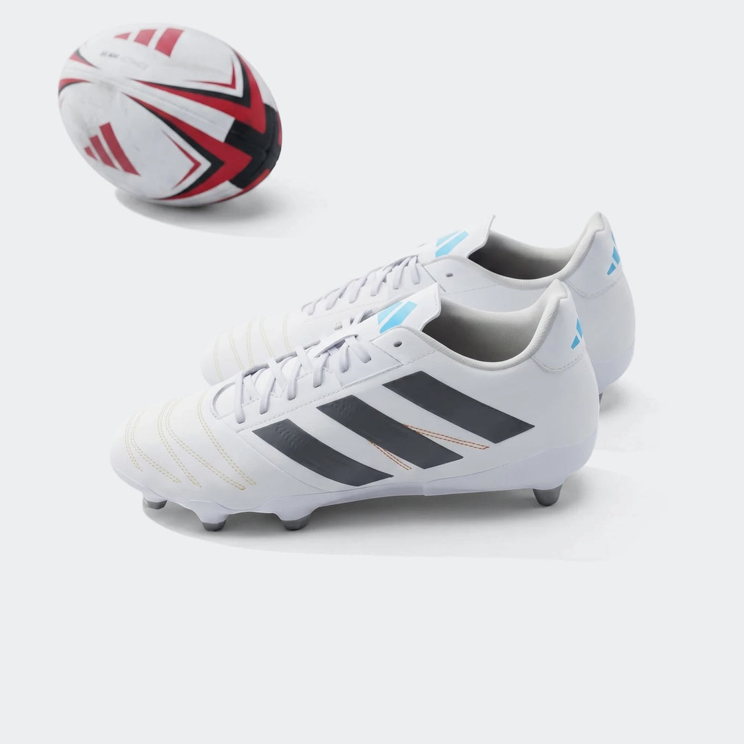 Pair of white Adidas Kakari Elite SG Rugby Boots with black stripes on a green grass field next to a rugby ball.