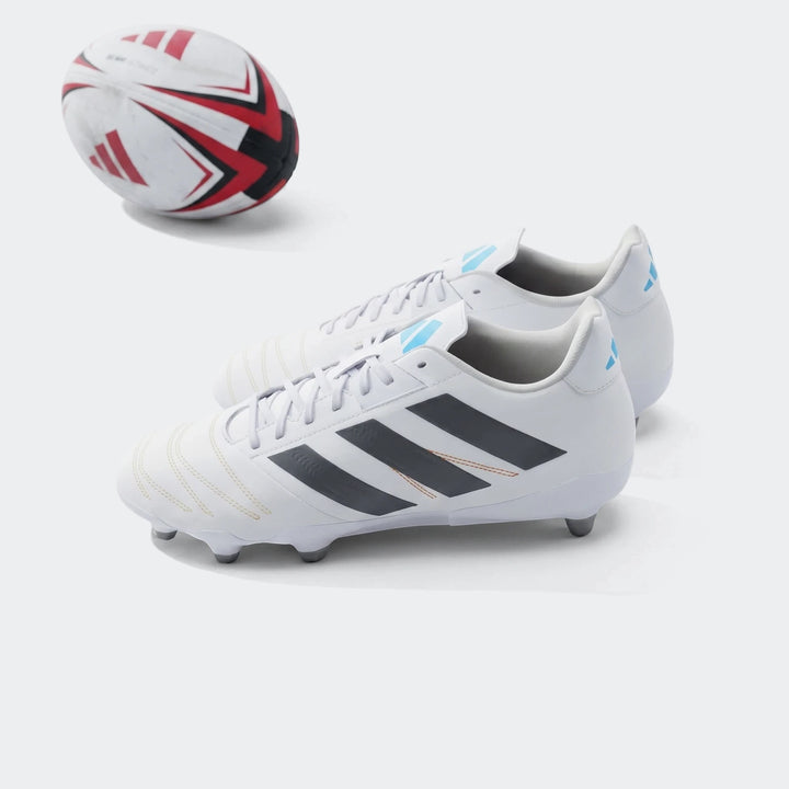 Pair of white Adidas Kakari Elite SG Rugby Boots with black stripes on a green grass field next to a rugby ball.
