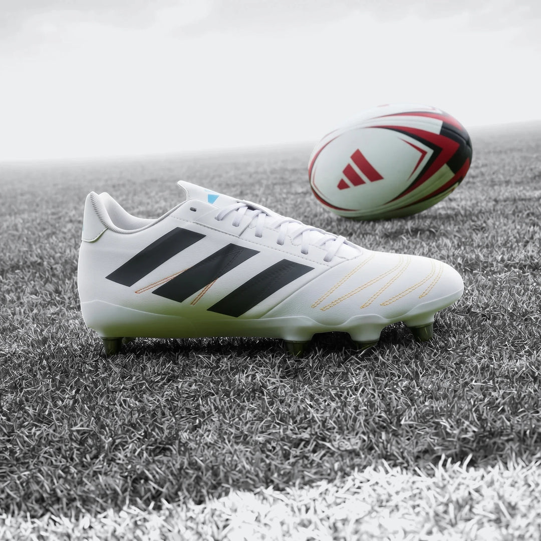 White Adidas Kakari Elite SG Rugby Boots with black stripes and orange stitching, placed on a green grass field next to a