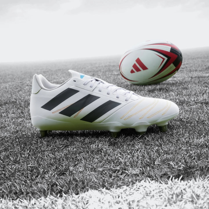 White Adidas Kakari Elite SG Rugby Boots with black stripes and orange stitching, placed on a green grass field next to a