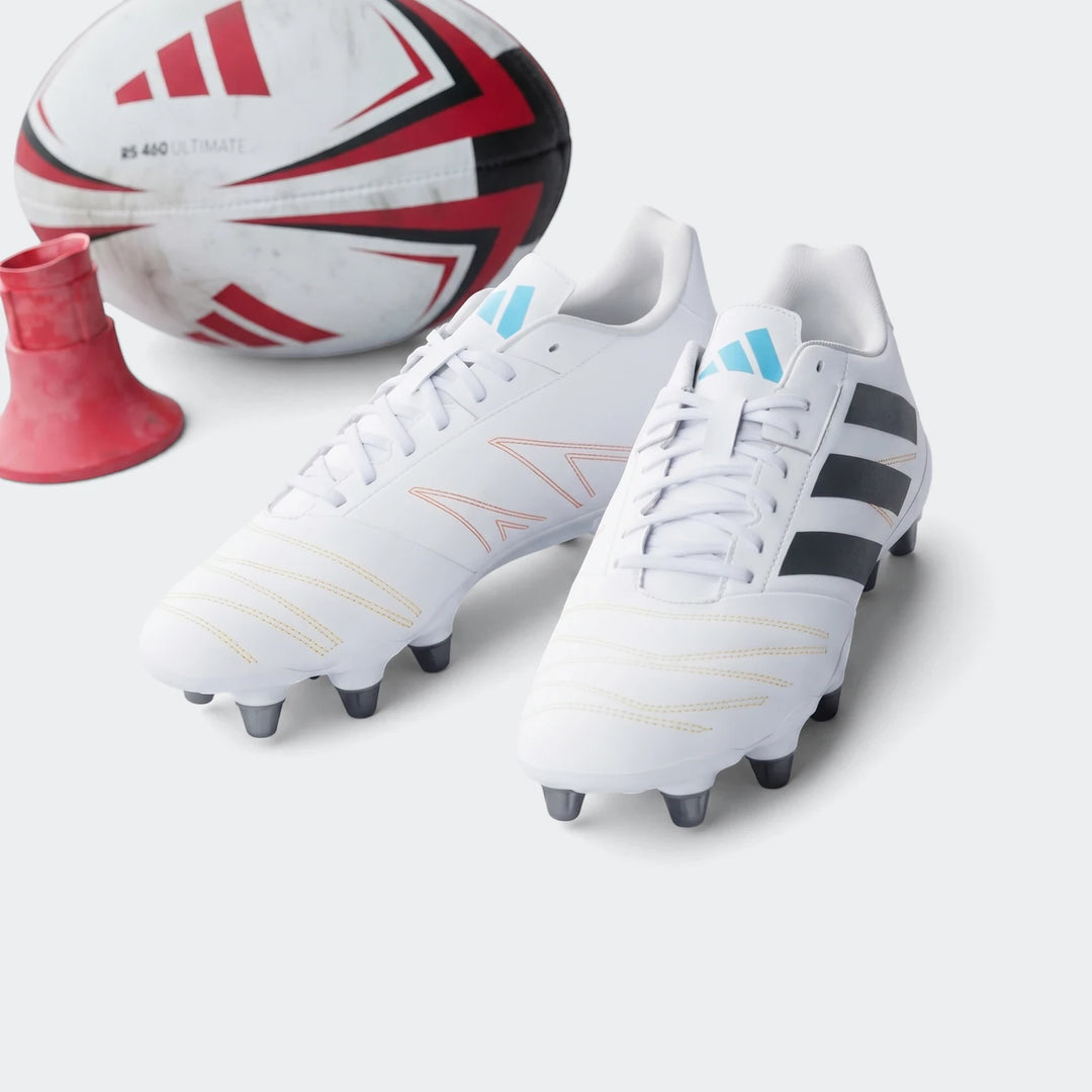 White Adidas Kakari Elite SG rugby boots with black stripes and blue logos, on a green grass field with a rugby ball and