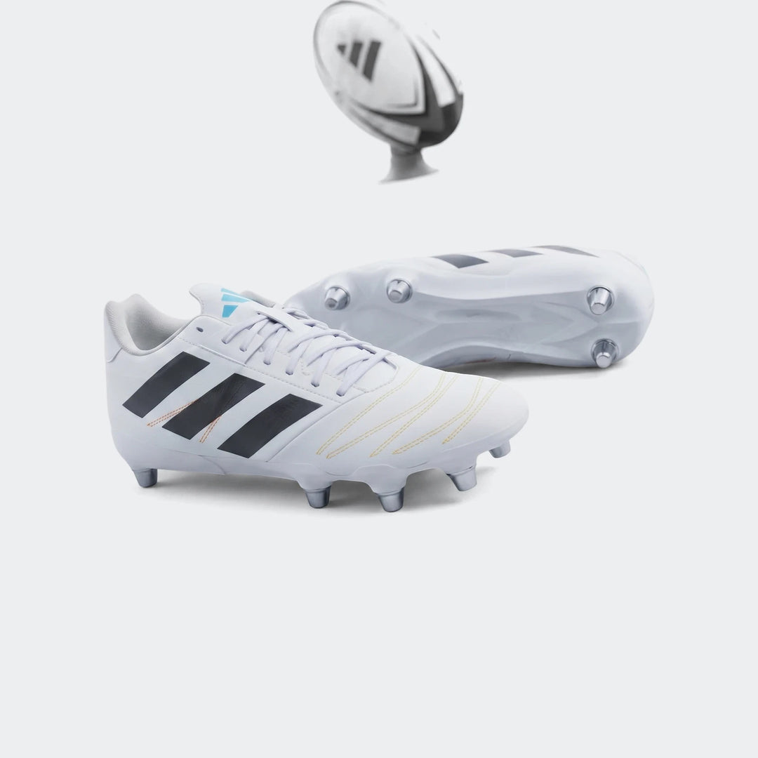 Pair of white Adidas Kakari Elite SG rugby boots with black stripes and blue accents, on a green grass field with a rugby