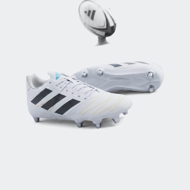 Pair of white Adidas Kakari Elite SG rugby boots with black stripes and blue accents, on a green grass field with a rugby