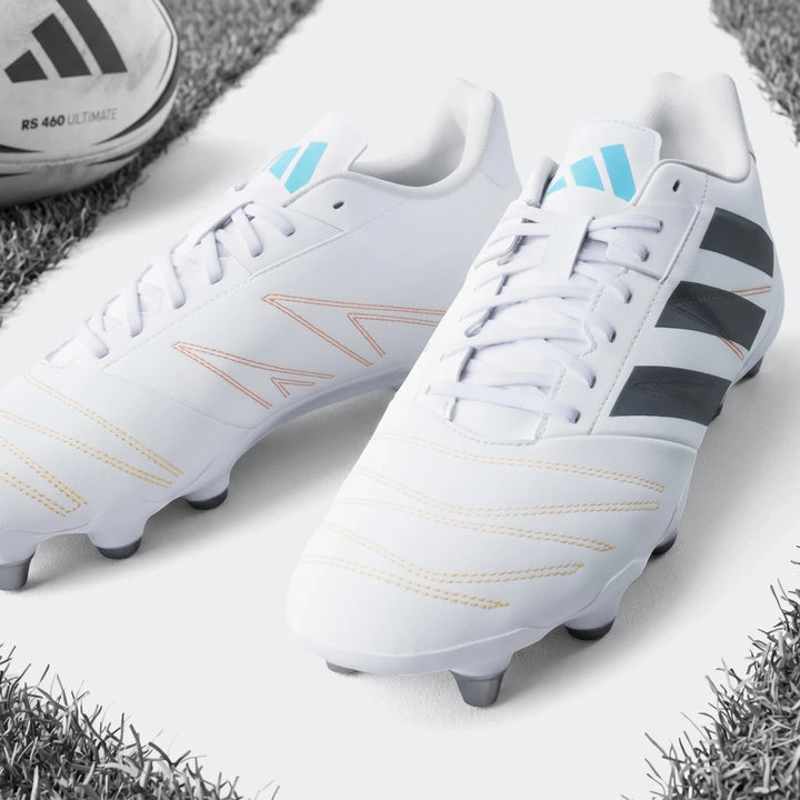 White Adidas Kakari Elite SG Rugby Boots with blue and orange accents, shown on a green grass field with a rugby ball.