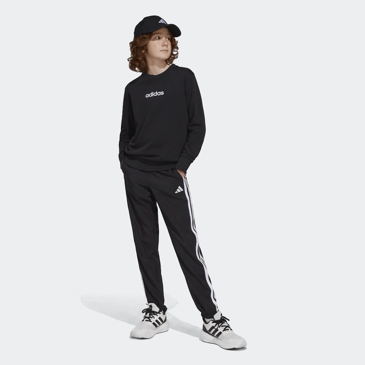 Young boy wearing black Adidas Essentials Climacool tracksuit bottoms and sweatshirt with a matching cap and white sneakers.